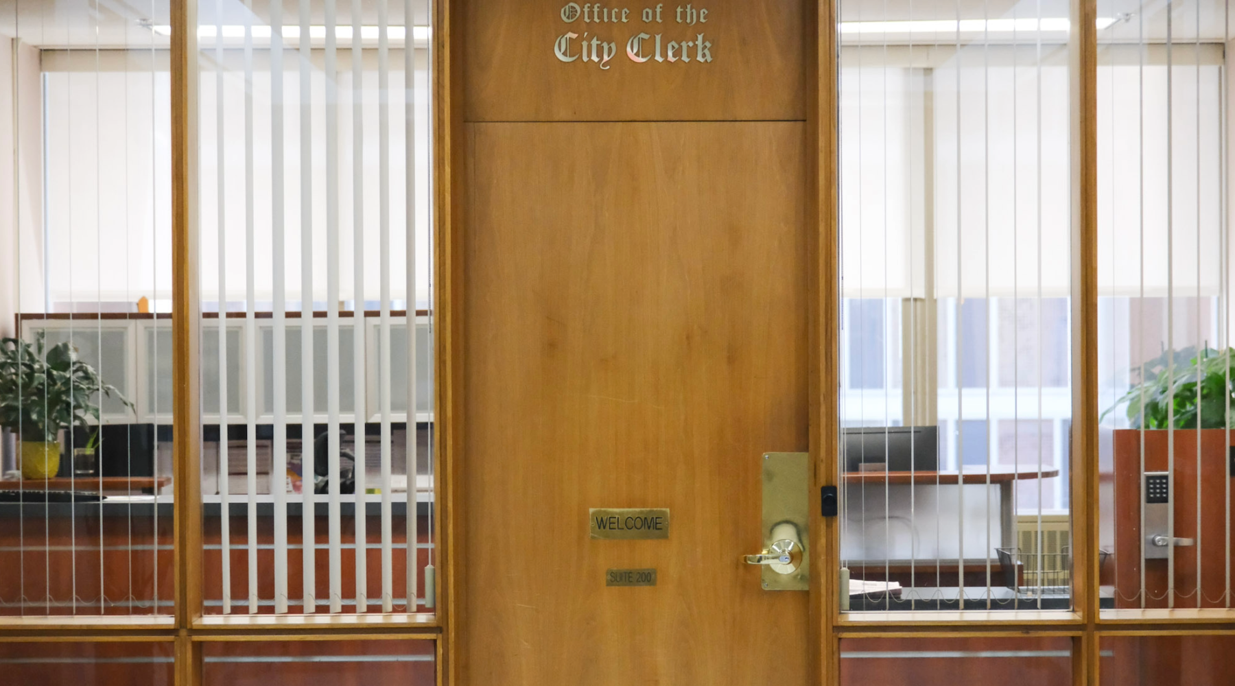 Wood-paneled door labeled “Office of the City Clerk,” centered in a glass wall with vertical blinds, revealing desks and plants inside.