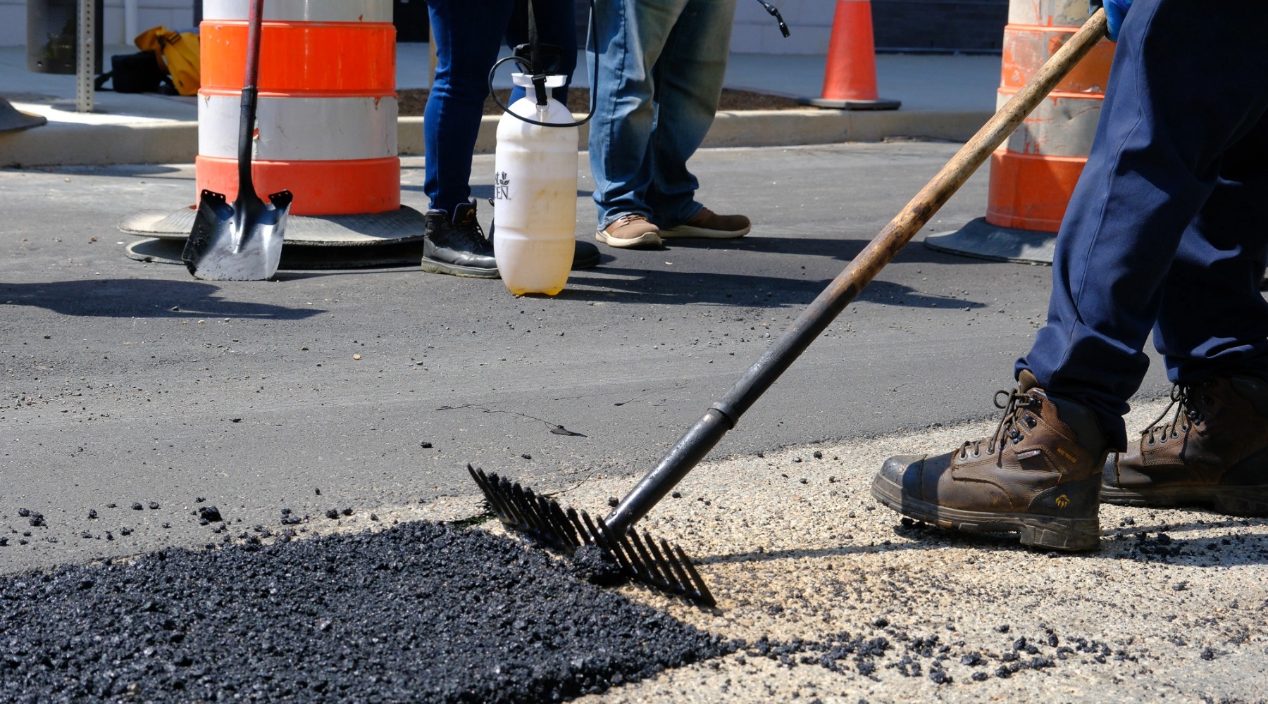 Crew member in work boots uses a rake to spread fresh asphalt during a street repair; traffic cones, shovels, and a spray tank in the background.