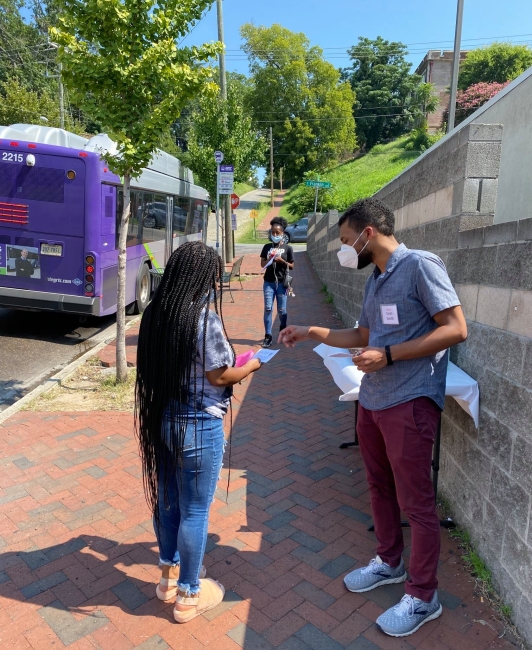 This image shows a person with short hair in a blue shirt administering a survey to a person with long braids outside by a purple bus.