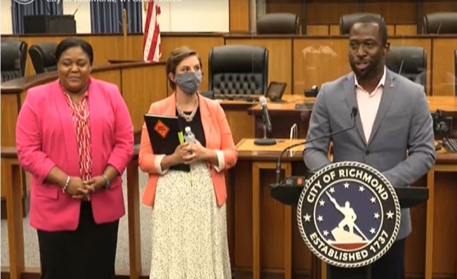 The image shows two business persons in pink blazers next to the Mayor who is standing at a podium with the City logo on front of the podium. 