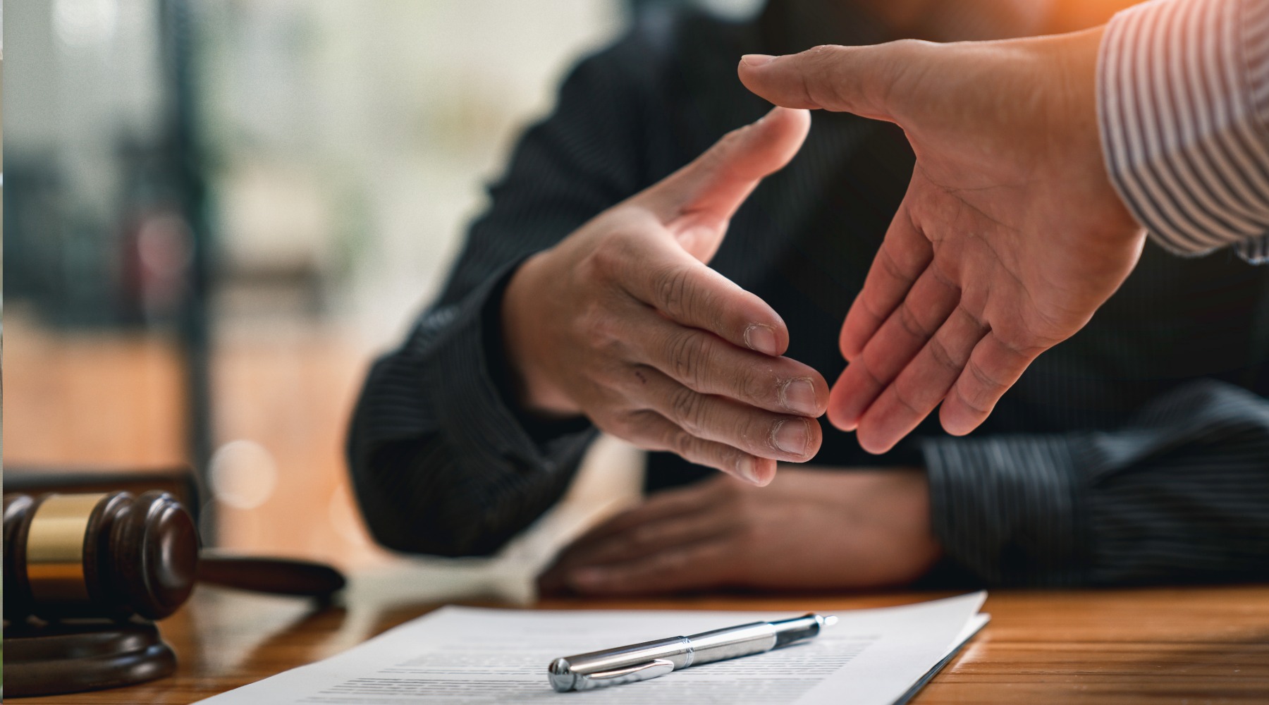 Two people extend hands for a handshake across a desk; a document and pen lie in the foreground with a gavel nearby, suggesting a professional or legal setting.