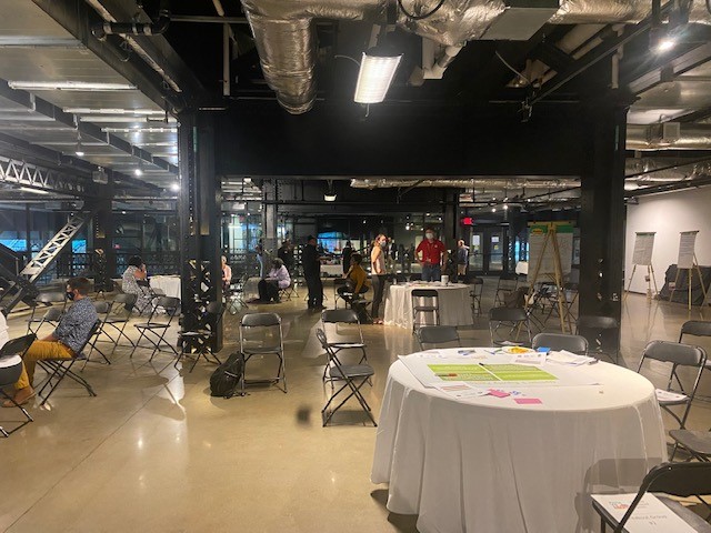 This image shows a meeting sapce set up with tables and chairs spaced far apart with people lingering in the background.