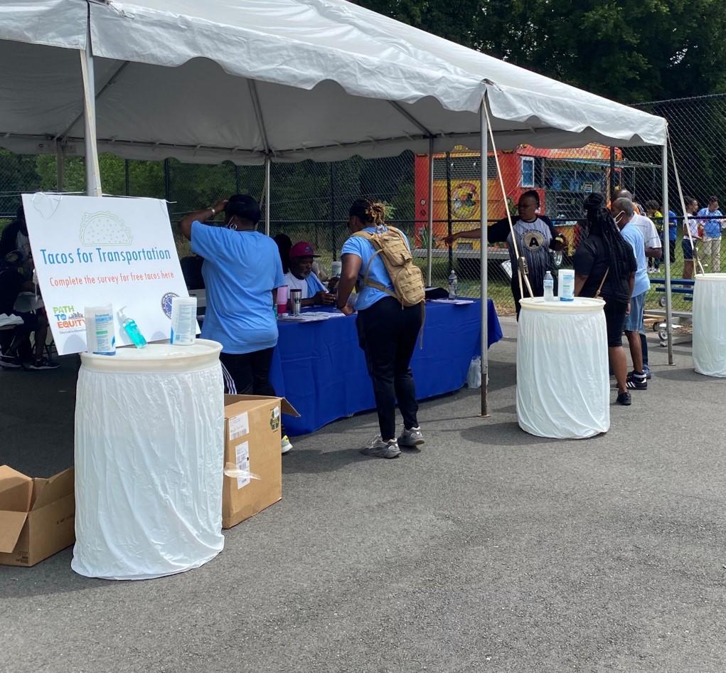 This image contains two people standing under an event tent with a taco truck in the baclkground and a community ambassador directing someone where to take a survey. 