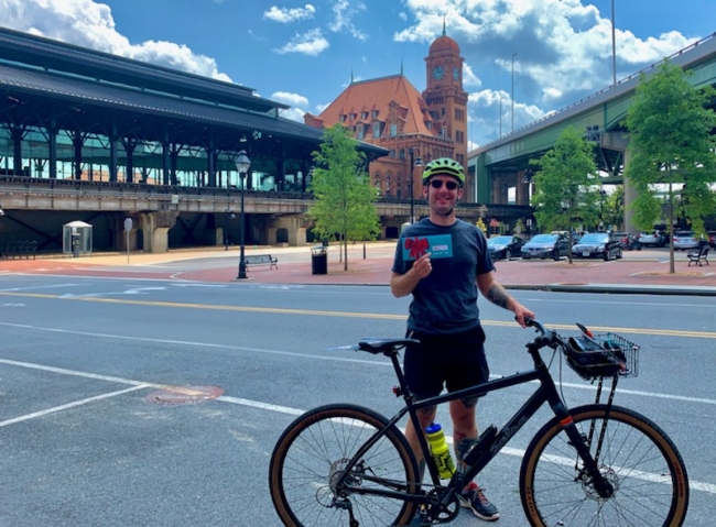 A person with a bike helmet holding a gift card envelope stands in from of main street station