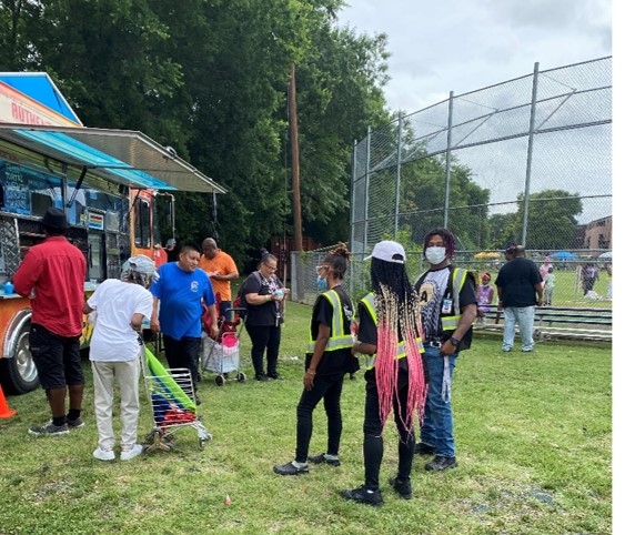 The image shows several people in line for a taco truck. Three people have yellow vests and are ready to administer surveys. 